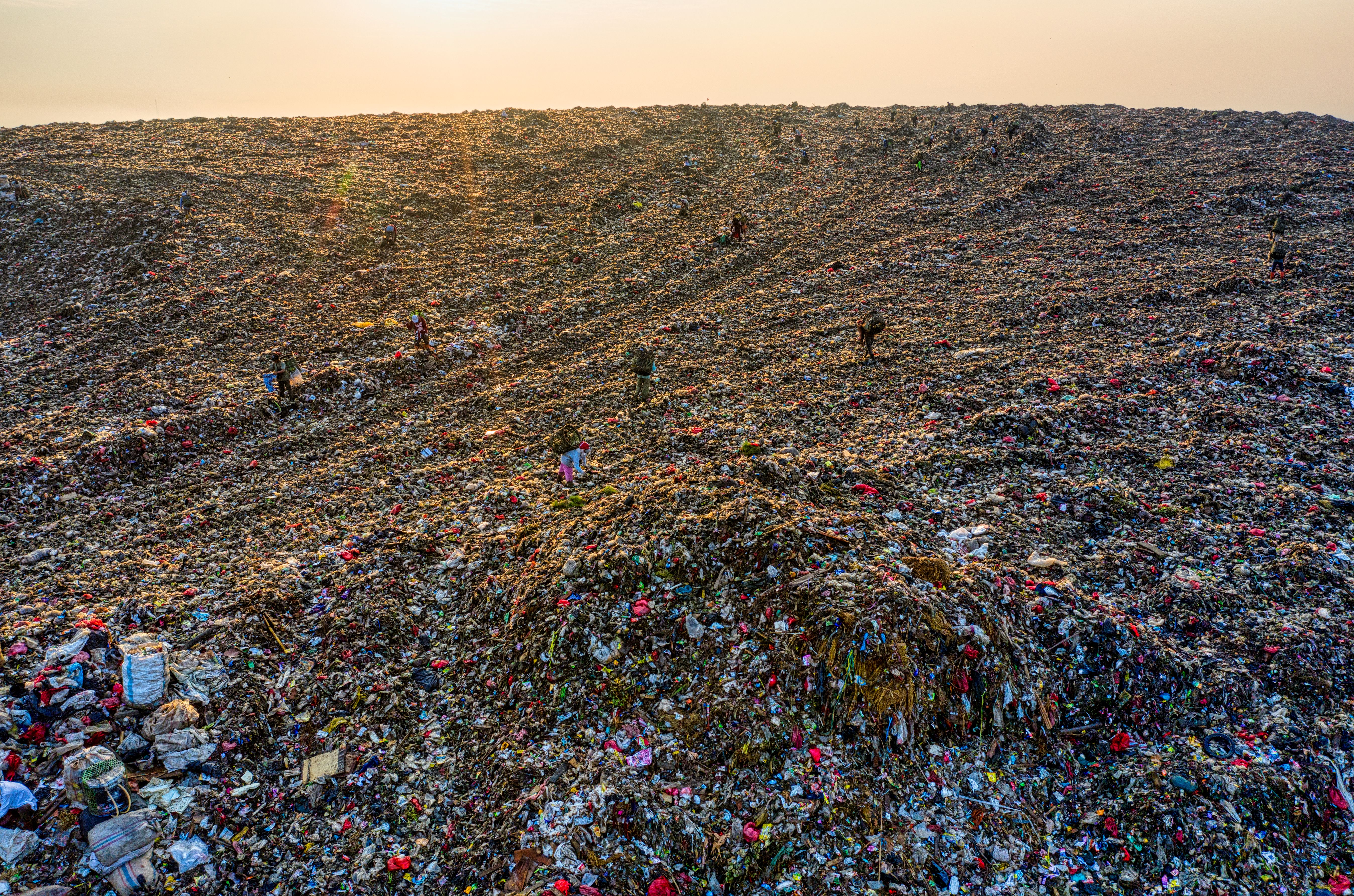 Aerial view of a vast landfill at sunset, tiny human figures across the surface