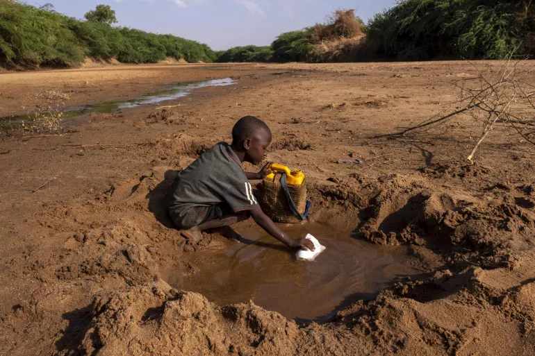 A young boy collecting muddy water from a hole in a dry riverbed, yellow jerrycan beside him